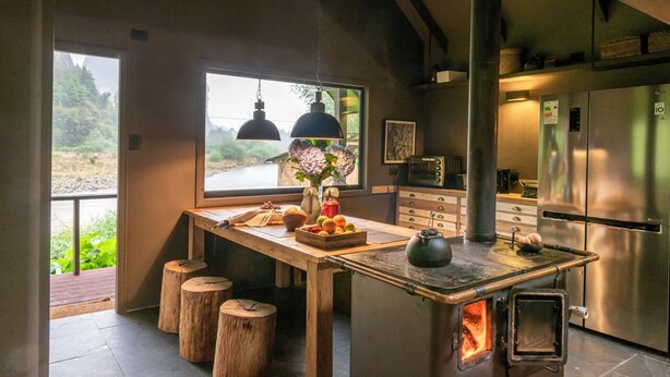 A modern kitchen with a wood-burning stove, stainless steel appliances, and a view of greenery outside. - Parque Cascadas, CABIN Rio Simpson-Patagonia Chile (Aysén)