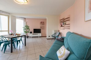 Ein Wohnzimmer mit einem türkisfarbenen Sofa, einem Esstisch mit Stühlen, einem Fernseher und einer Pflanze.