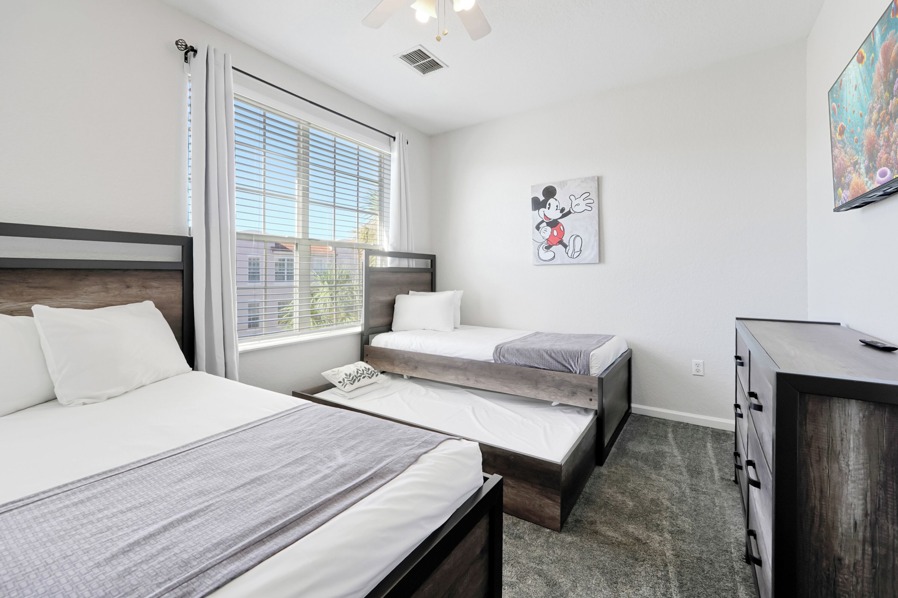 A modern hotel room with two beds, a large window with blinds, a ceiling fan, a wall-mounted TV, and a wooden dresser.