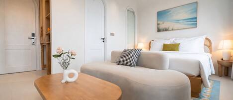 A modern hotel room with a curved sofa, a wooden coffee table, a painting of a beach, and a bed with white bedding.