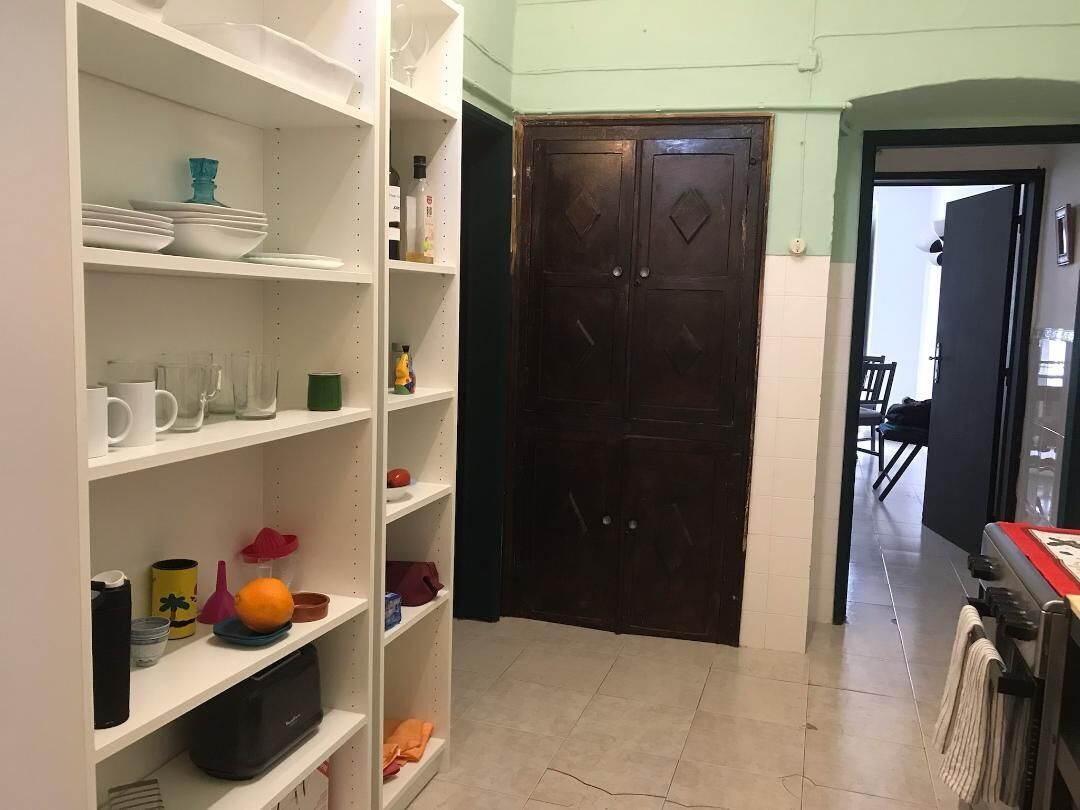 A kitchen with white shelves, a dark wooden door, and a dining area visible through an open door.