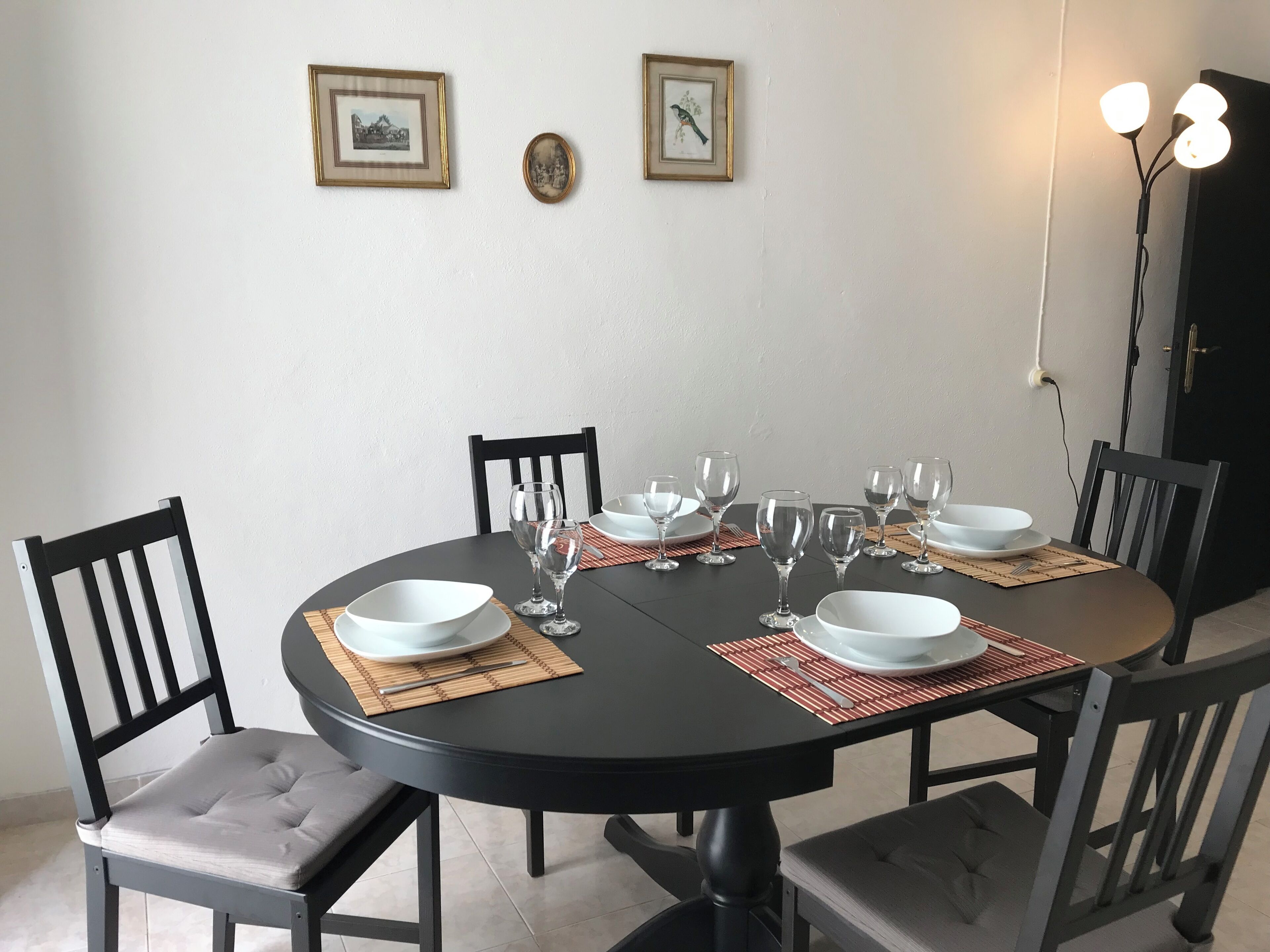 A round dining table with six chairs, each with a cushioned seat and backrest. The table is set with white plates, clear wine glasses, and red placemats. Two framed pictures hang on the wall above the table.