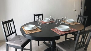 Una mesa redonda de comedor con seis sillas, cada una con asiento y respaldo acolchados. La mesa está dispuesta con platos blancos, copas de vino transparentes y salvamanteles rojos. Dos cuadros enmarcados cuelgan de la pared sobre la mesa.