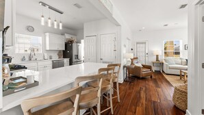 Un'ampia cucina con mobili bianchi, un'isola grande e sedie in legno. C'è una zona pranzo con tavolo e sedie, e un soggiorno con divano e lampada.