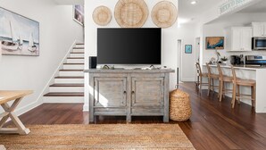 Un soggiorno moderno con una TV a schermo piatto, una scala e un angolo cottura con pensili bianchi e sedie di legno.
