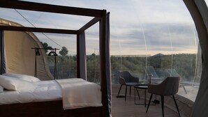 A bedroom with a bed, a table, and two chairs, a large window with a view of trees and a bridge, and a tent-like structure. - Panoramic Suites (Guadamur)