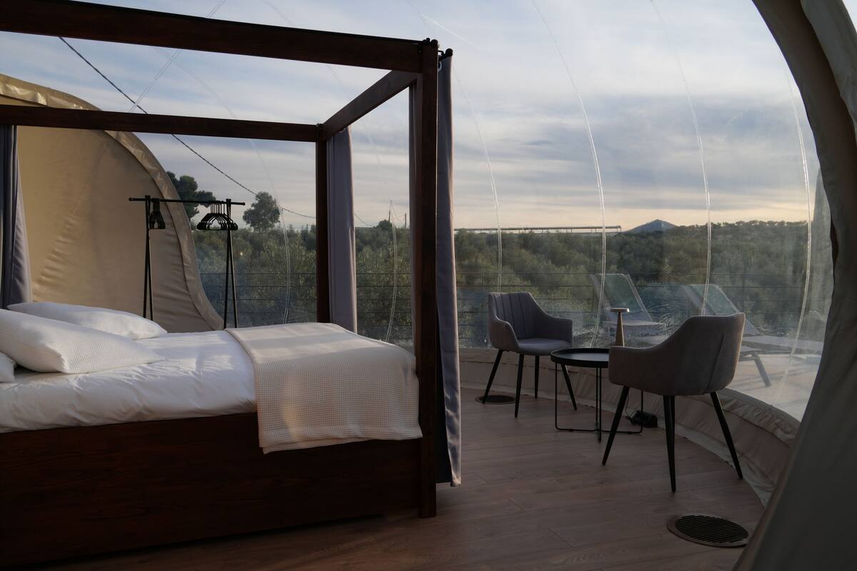 A bedroom with a bed, a table, and two chairs, a large window with a view of trees and a bridge, and a tent-like structure.