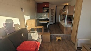 Un espace de vie compact comprenant un coin cuisine, un canapé avec un coussin rouge, une table en bois et un lit superposé.