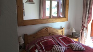 Un dormitorio con cama de madera, un espejo grande, una mesita de noche con lámpara y una ventana con cortinas.