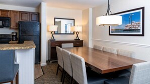 Une chambre d’hôtel dotée d’un coin repas avec une table et des chaises en bois, d’une cuisine équipée d’un réfrigérateur et d’un four à micro-ondes, d’un grand miroir et d’un tableau encadré au mur.