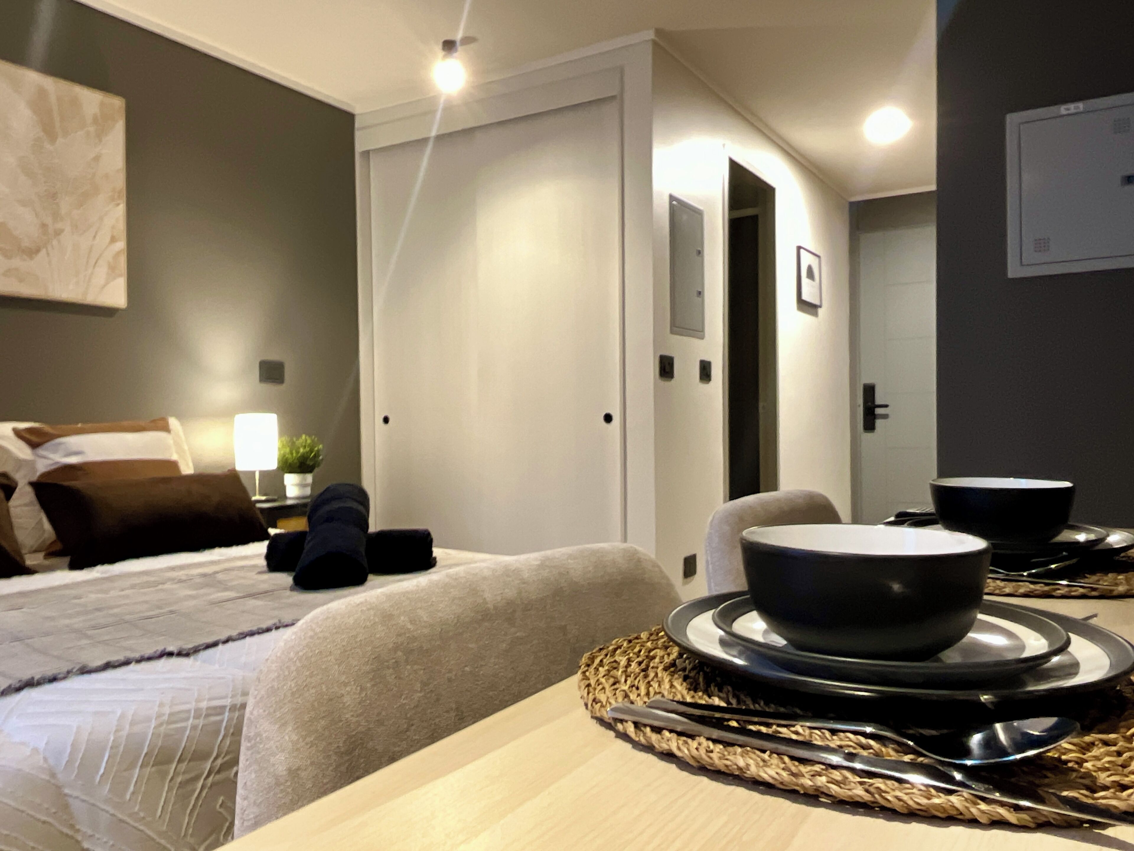 A modern hotel room with a neatly made bed, a dining table set with plates and cutlery, and a small potted plant on the bedside table.