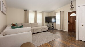 A living room with a beige sofa, a flat-screen TV on a stand, a wooden floor, and a door with a glass panel. - Beautifully furnished house located in sought after Rosedale community. (Rosedale)