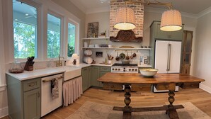 Una cocina con una mesa de madera, estufa y refrigerador. Hay ventanas, un fregadero y una encimera con varios objetos.