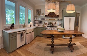 A kitchen with a wooden table, a stove, and a refrigerator. There are windows, a sink, and a countertop with various items.
