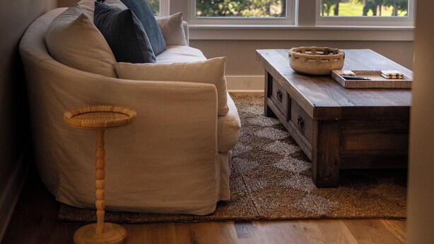 Una habitación acogedora con sofá, lámpara, mesa de centro de madera y vistas a árboles a través de amplias ventanas.