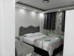 A modern bedroom with a grey tufted headboard, a grey sofa, a patterned bedspread, and a brick wall accent. - Nexus By Taksim Hotel  (İstanbul)