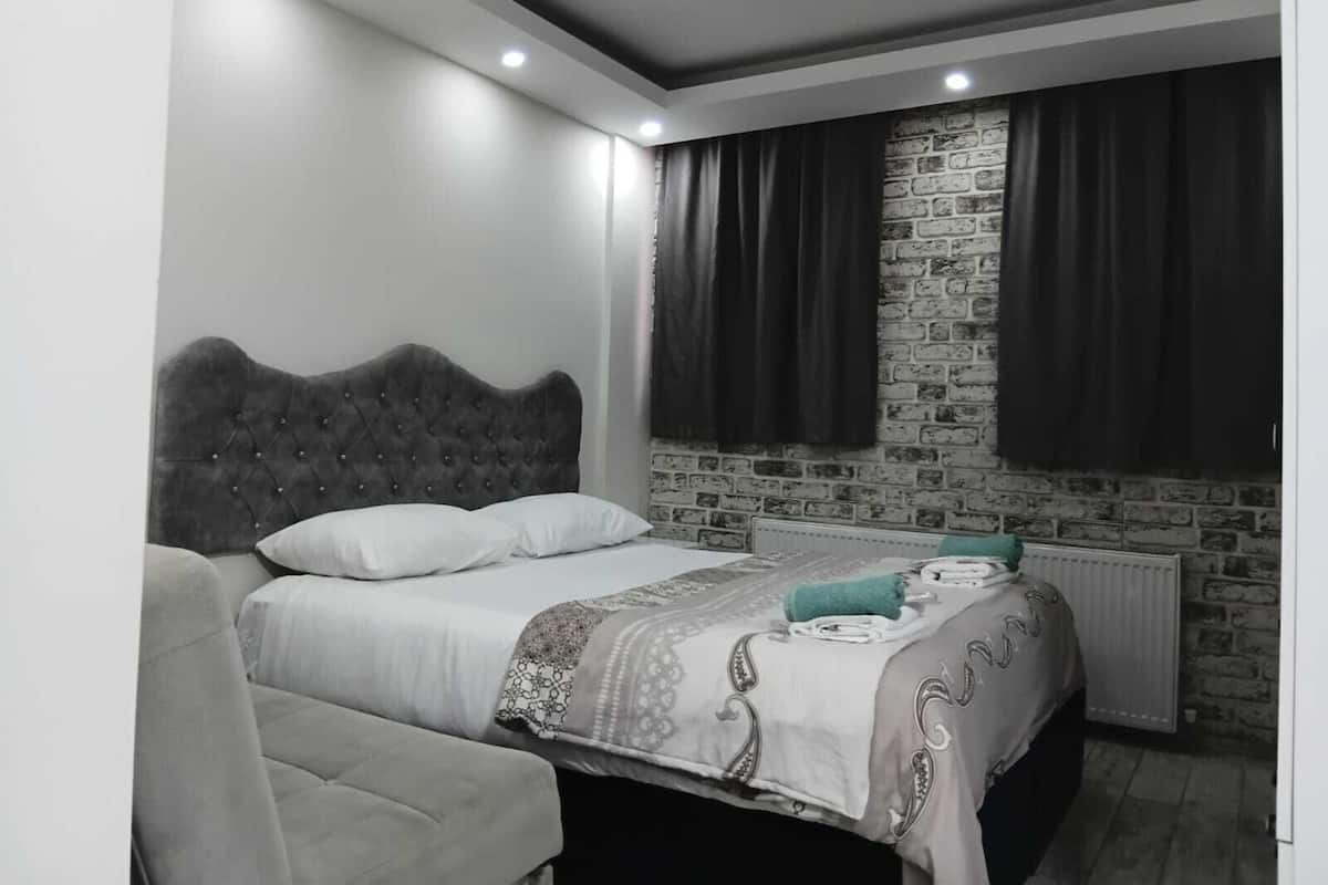 A bedroom with a grey tufted headboard bed, white bedding, a grey sofa, a brick-patterned wall, and a radiator.
