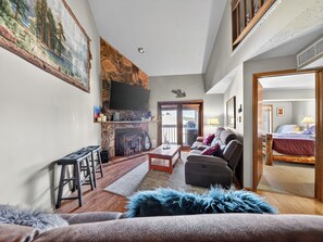Ein Wohnzimmer mit Kamin, einem an der Wand montierten Fernseher, einer Couch, einem Couchtisch und Blick durch eine Glastür ins Freie.