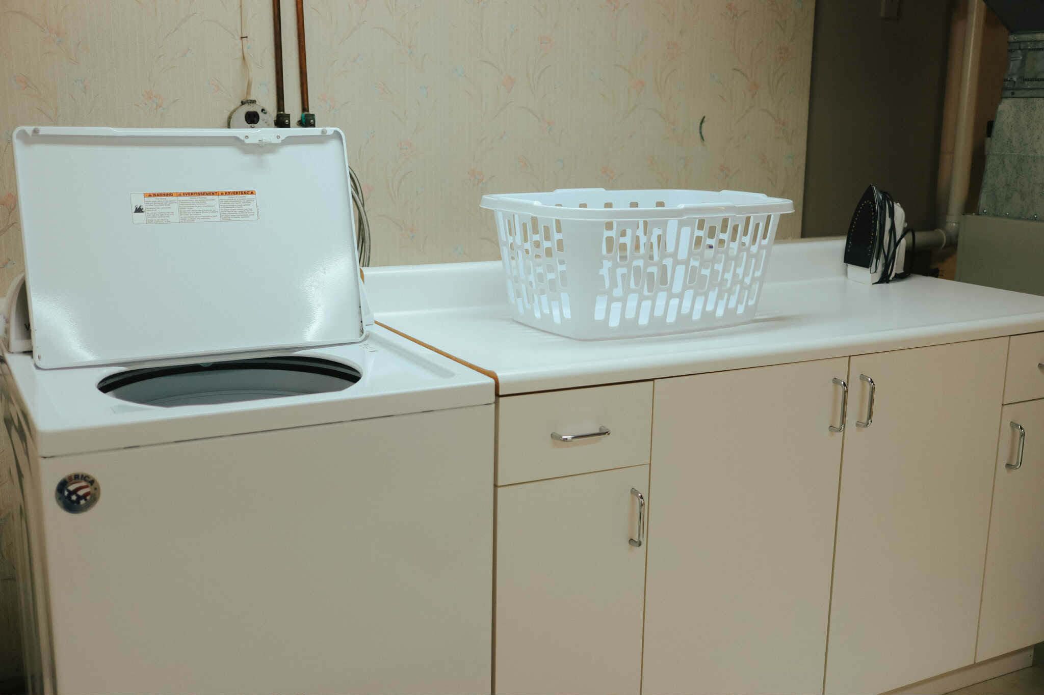 Una lavadora blanca con una cesta y un electrodoméstico pequeño sobre una encimera blanca.