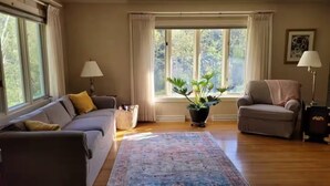 Ein Wohnzimmer mit grauem Sofa, einem beigen Sessel, einem großen Fenster mit Vorhängen, einer Topfpflanze und einem gemusterten Teppich.