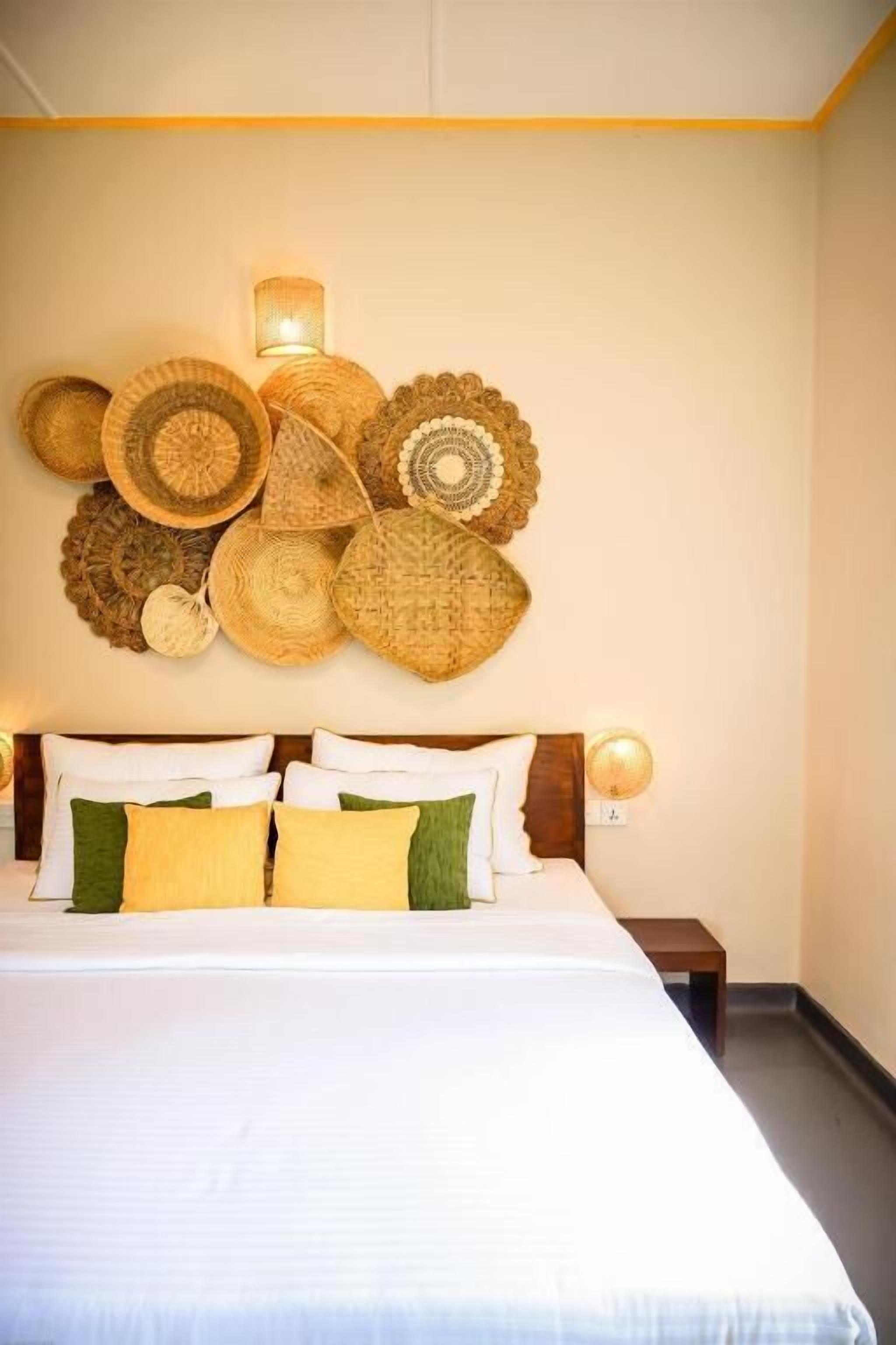 A neatly made bed with white and green pillows, a wooden headboard, and a wall decorated with woven baskets and a wall-mounted lamp.