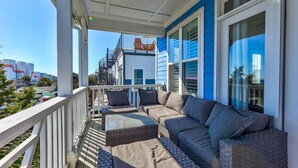 A covered porch with a sofa and chairs, a railing, and a view of a building with a blue exterior and a red sign. - Hightide Oasis is a bright, coastal getaway short walk to the beach. (Carolina Beach)