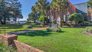 A church with a red roof and wooden exterior, surrounded by well-maintained grass and palm trees. - Two Homes · Walk to Old Town · Fire Pit · EV (Bluffton)