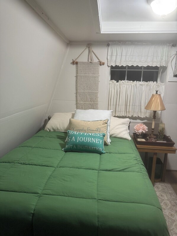 Une chambre avec une couvre-lit verte, deux oreillers, une décoration murale en macramé, une fenêtre avec des rideaux blancs, une lampe de chevet et une petite table avec un plateau.