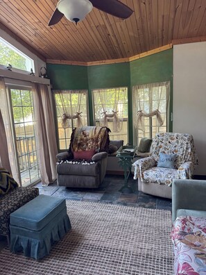 A cozy room with wooden ceiling, green walls, two windows with curtains, a sofa, a chair, and a small table.