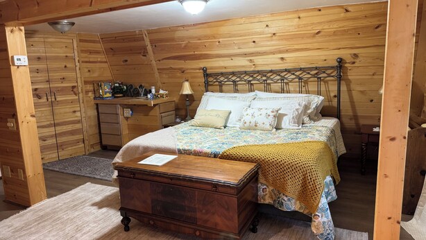 A wooden cabin bedroom with a bed, dresser, nightstand, lamp, and a small desk with items on it.
