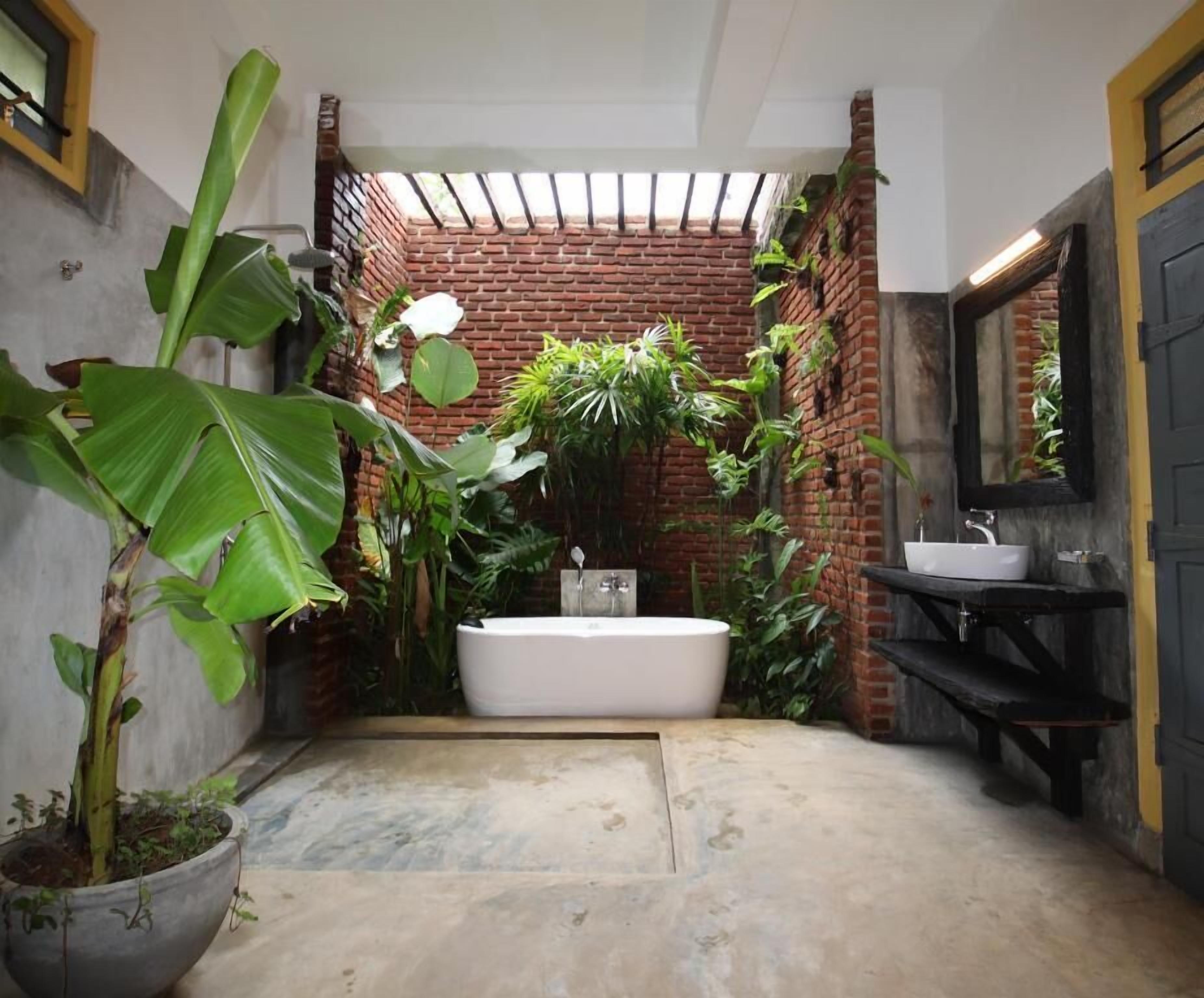 Een badkamer met een bakstenen muur, een grote potplant, een witte badkuip en een zwarte wastafel.