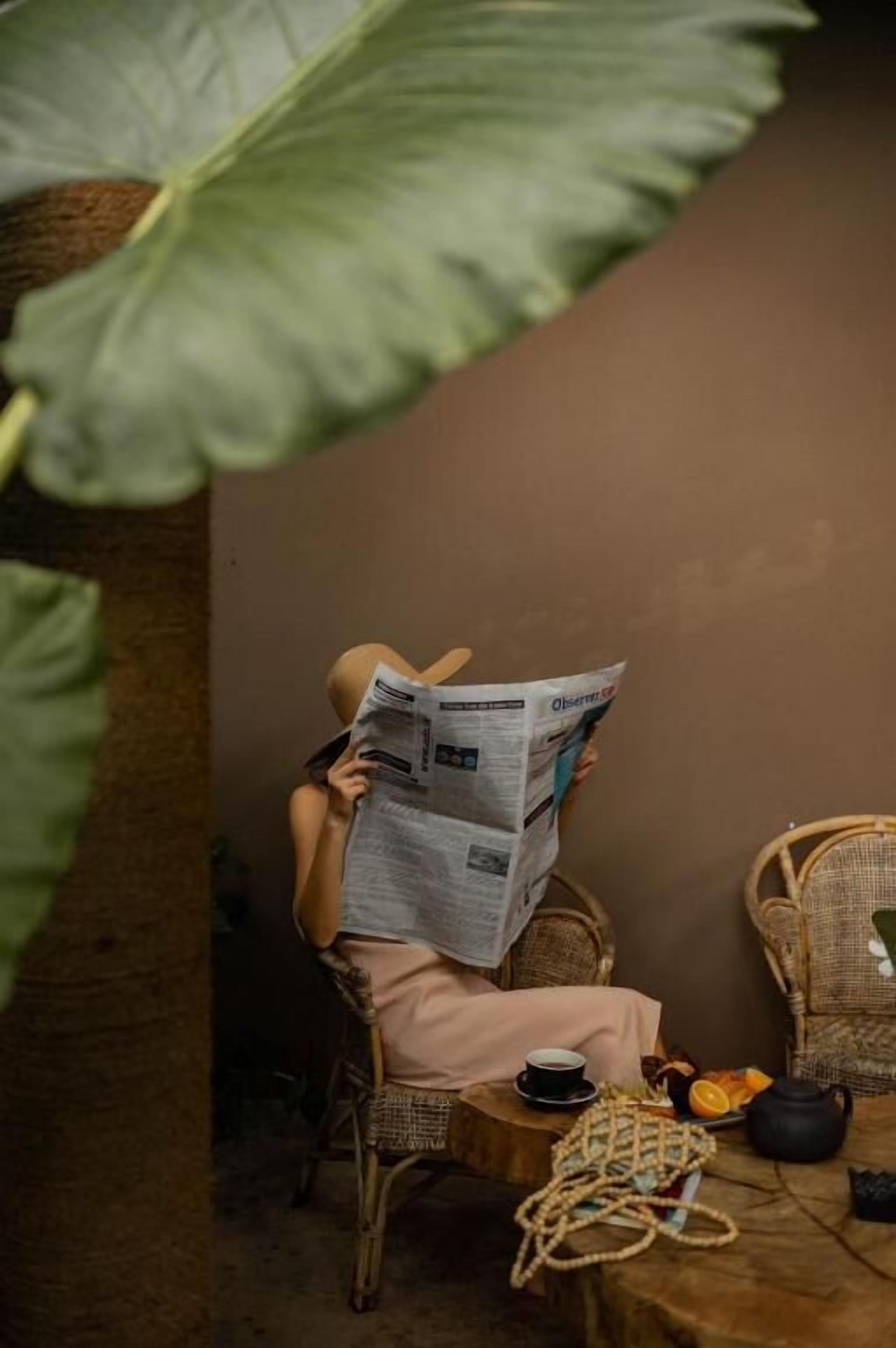 A person sitting on a wicker chair in a room with a large green plant, reading a newspaper. There is a small table with a teapot, a cup, and some fruit.