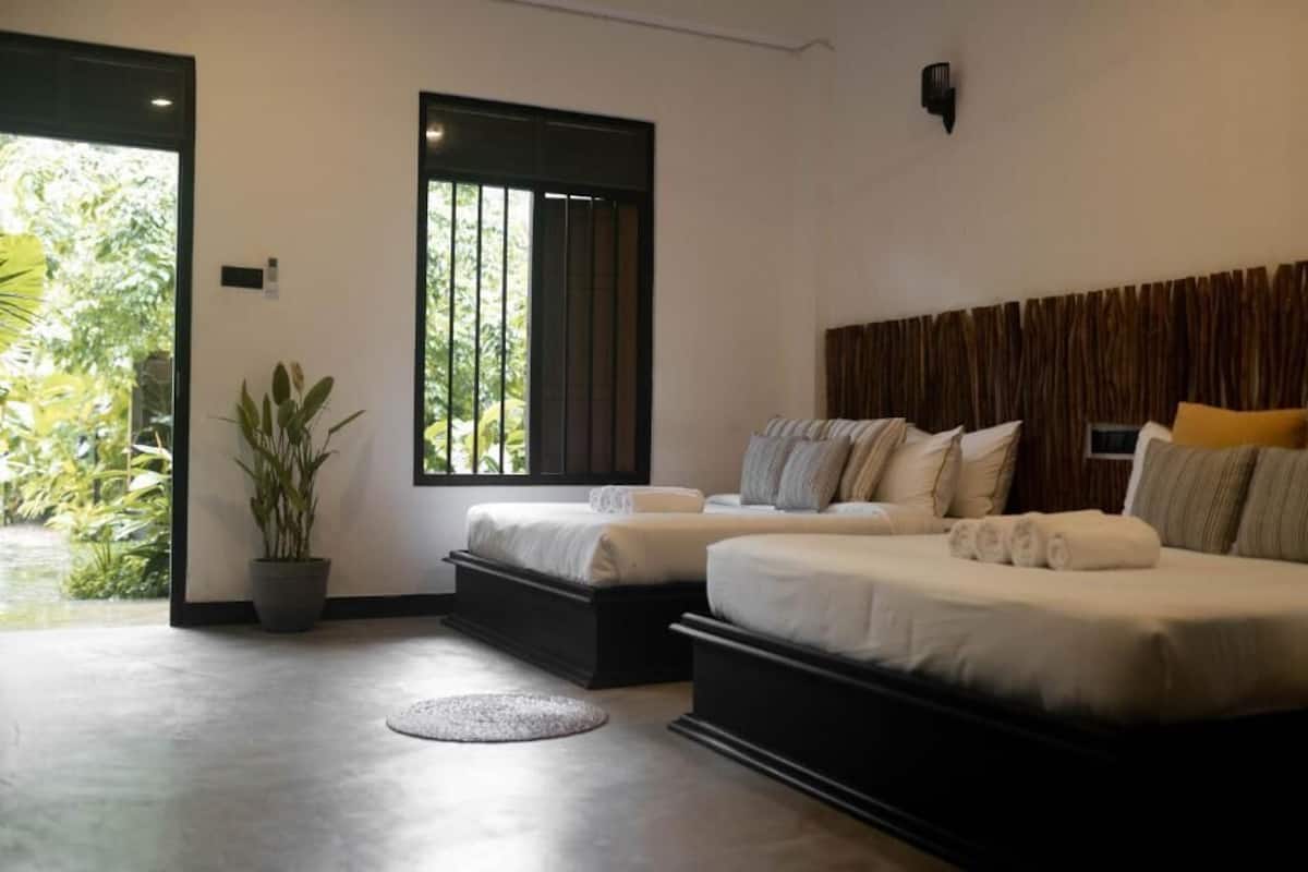 A modern bedroom with two beds, a potted plant, and a view of greenery outside.