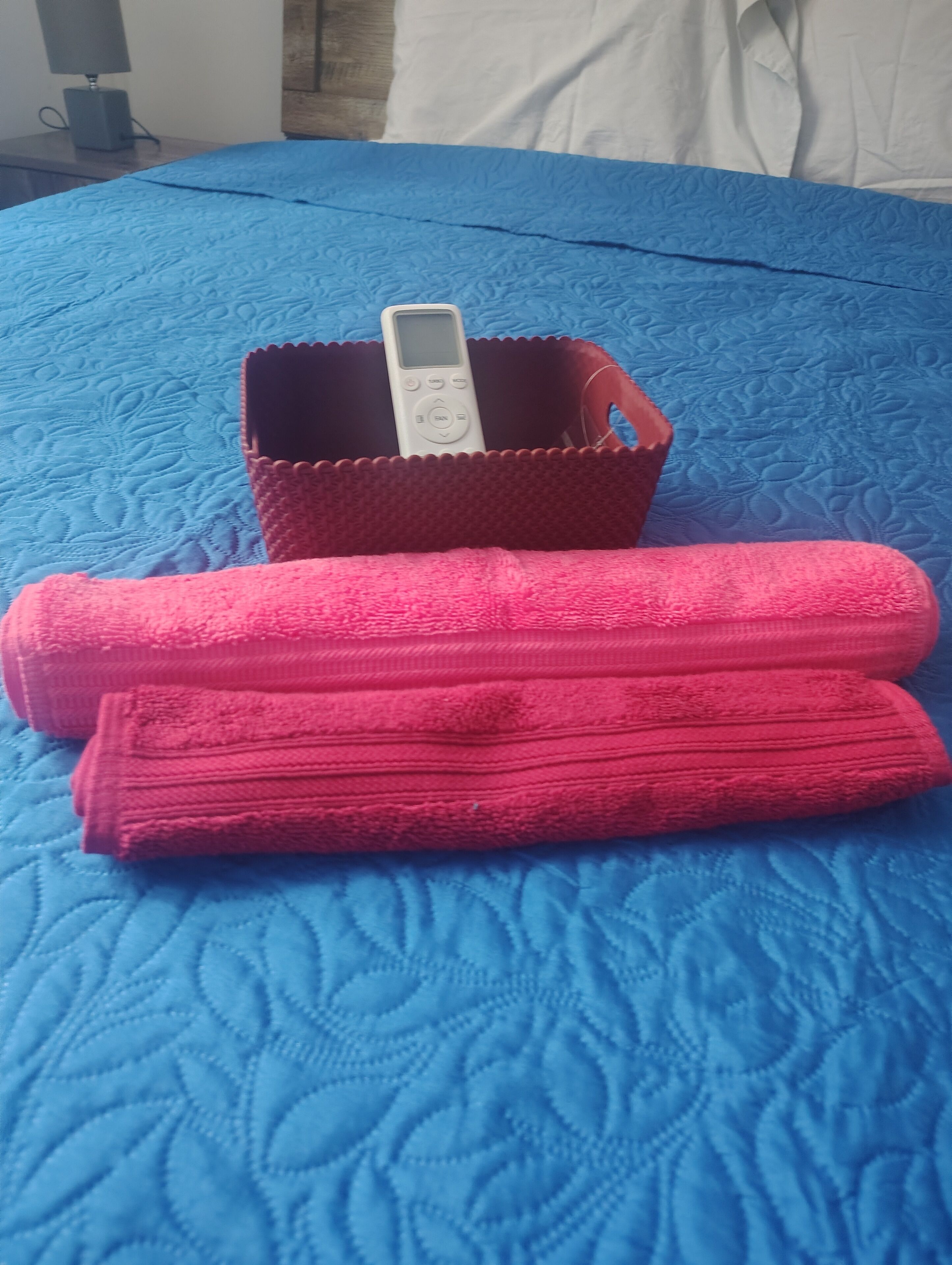 Un lit avec une couette bleue, un panier marron contenant un appareil blanc, et deux serviettes roses.