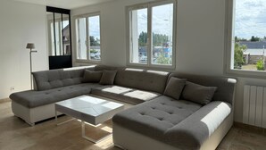 A modern living room with a sectional sofa, a small white table, and large windows offering views of the outside. - Spacious and Bright New T3 (Évreux)