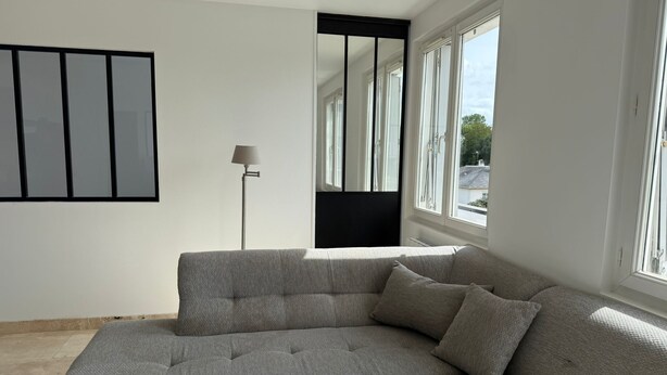 A modern living room with a grey sectional sofa, a floor lamp, and large windows offering a view of the outside. - Spacious and Bright New T3 (Évreux)