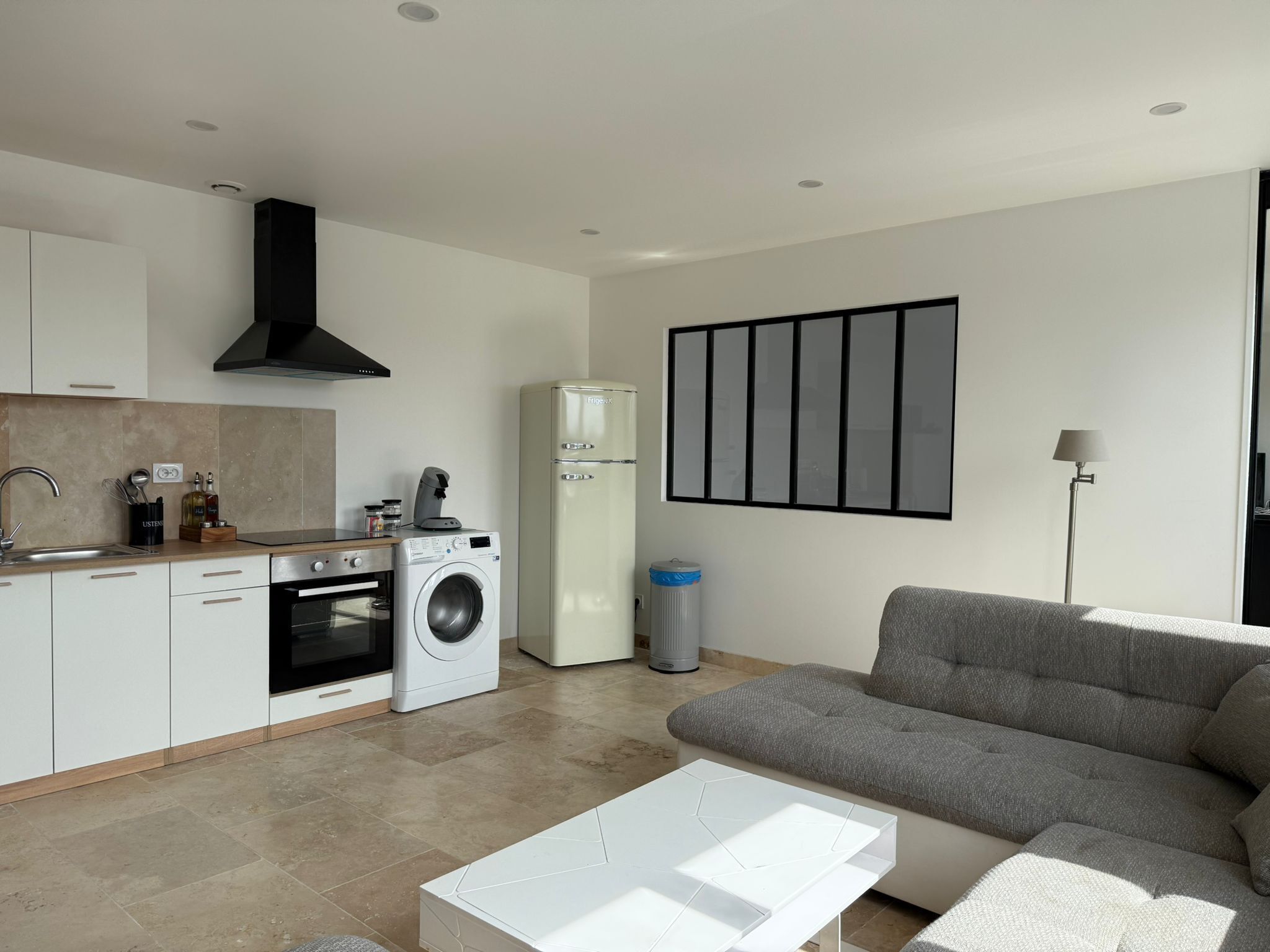 Een moderne keuken met een ingebouwde oven, een wasmachine en een koelkast. In de woonkamer staat een grijze bank en een witte salontafel.