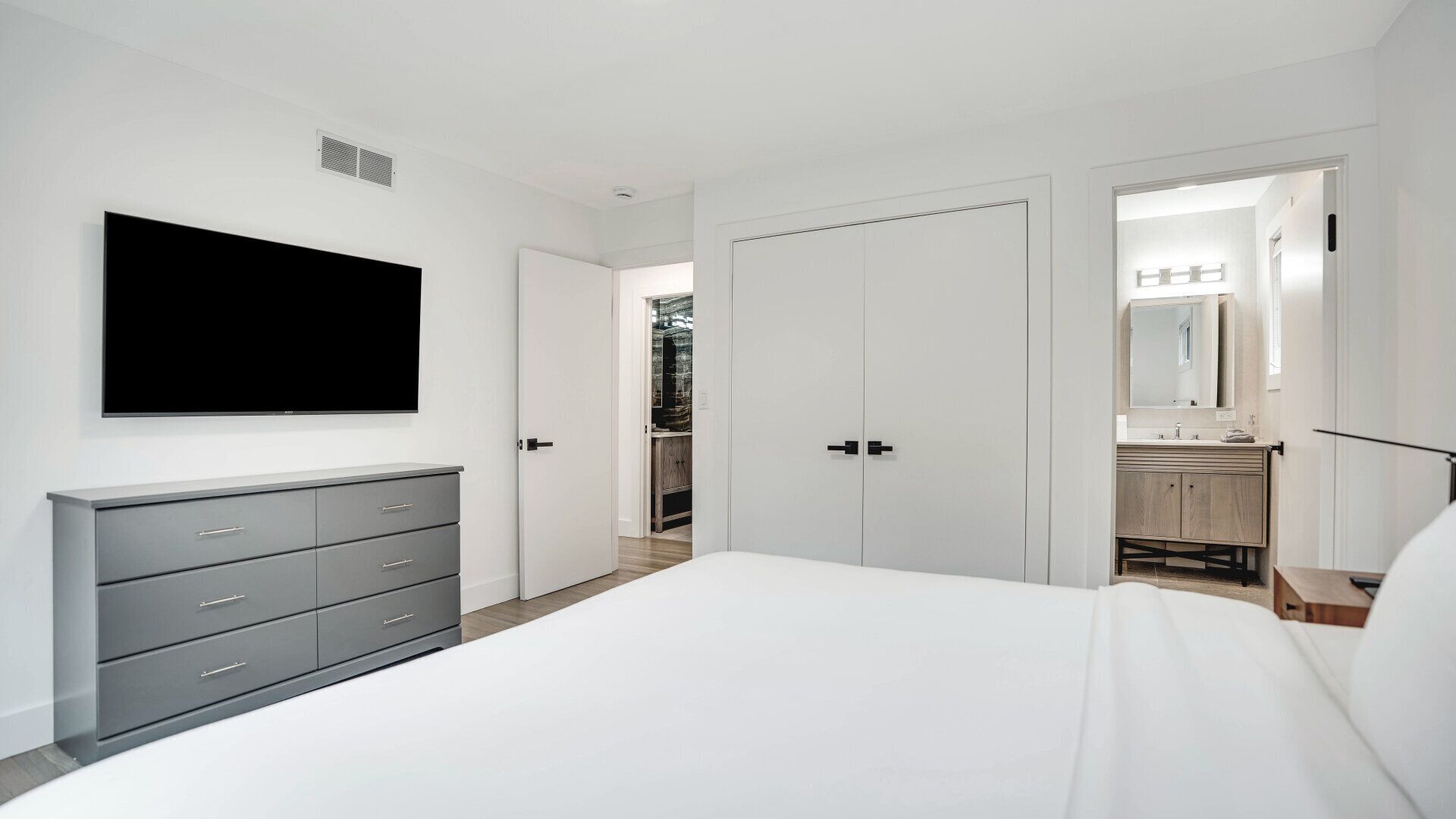 A bedroom with a bed, a dresser, a TV, and a bathroom visible through an open door.