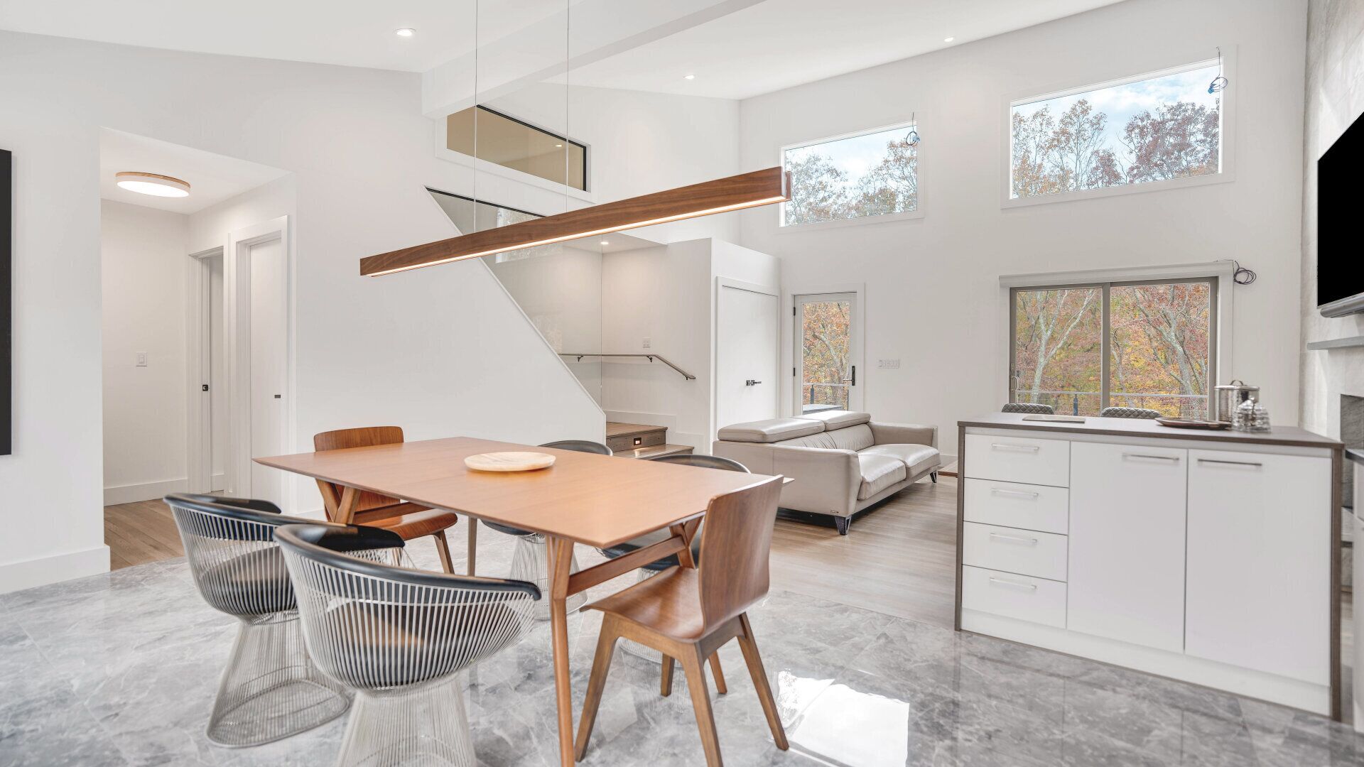 A modern kitchen with white cabinetry, a dining area with a wooden table and chairs, and a living area with a sofa and a TV.