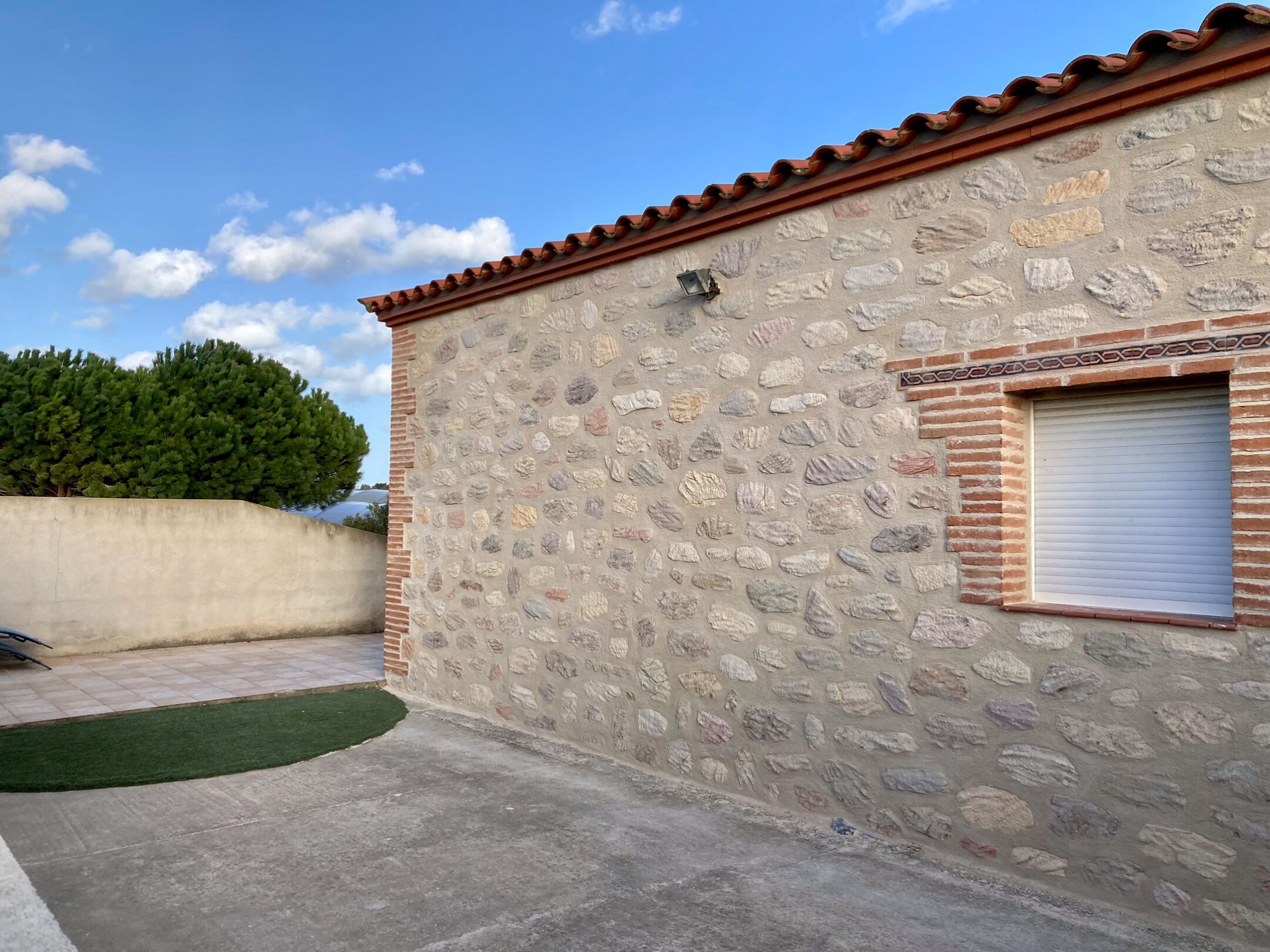 Un edificio in pietra con tetto in tegole, una piccola finestra con persiana e un muro in cemento con un prato verde di fronte.