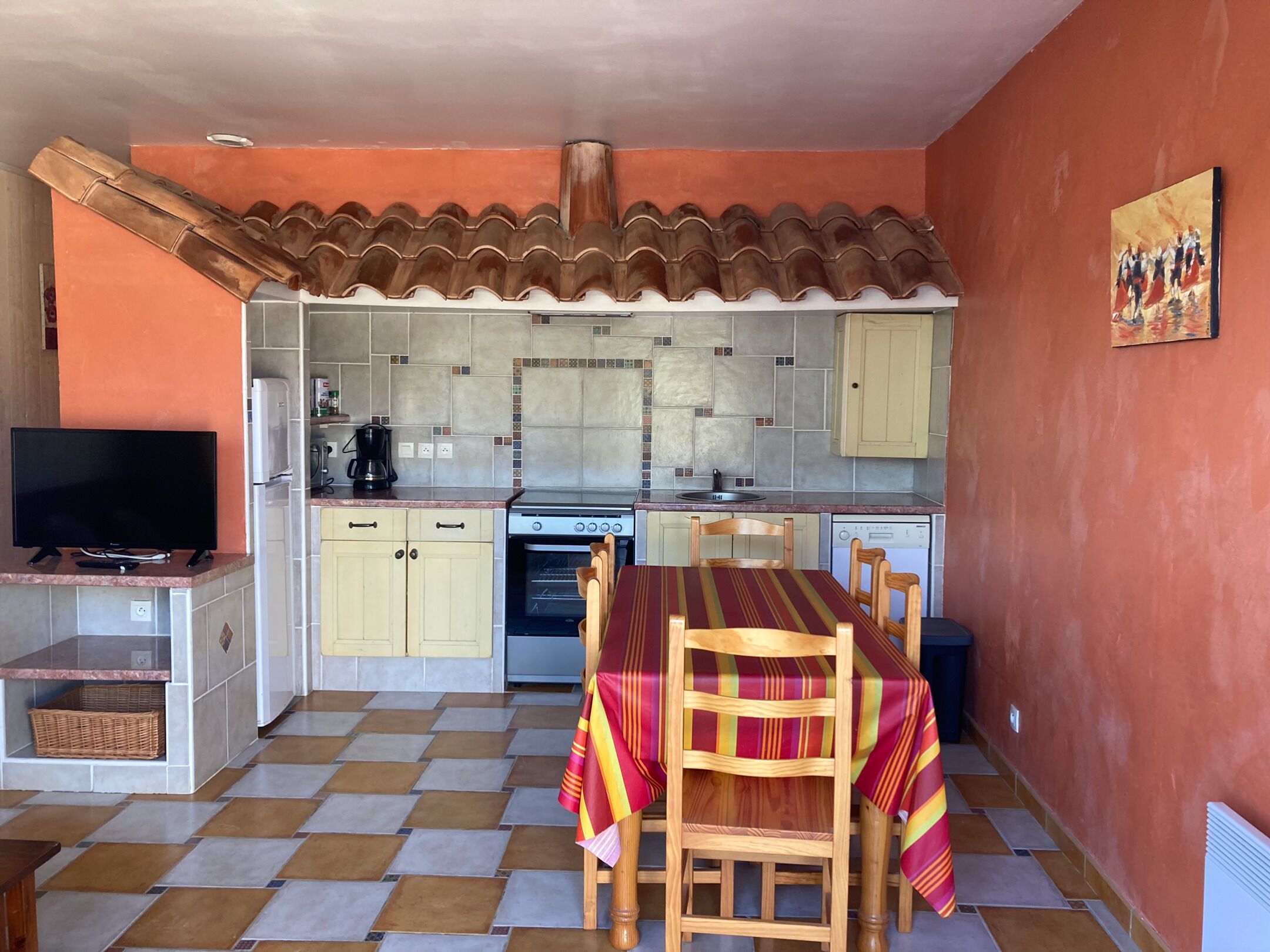 A compact kitchen with a tiled backsplash, a stove, and a dishwasher. There is a dining table with chairs and a television on a stand.
