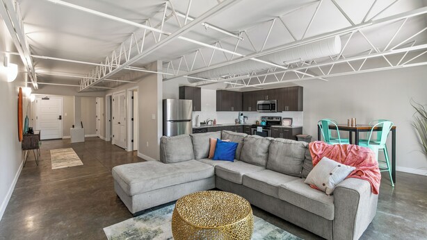 A modern living room with a grey sectional sofa, a round woven coffee table, and a kitchen area in the background. - Relax & Recharge 1br Downtown Loft w Private Patio (Des Moines)