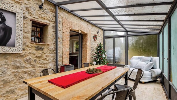 A dining area with a wooden table, chairs, and a red placemat. A white sofa is against a glass wall, and a Christmas tree is visible in the background. - Charming Tuscany Villa w/ Private Infinity Pool (Castel del Piano)