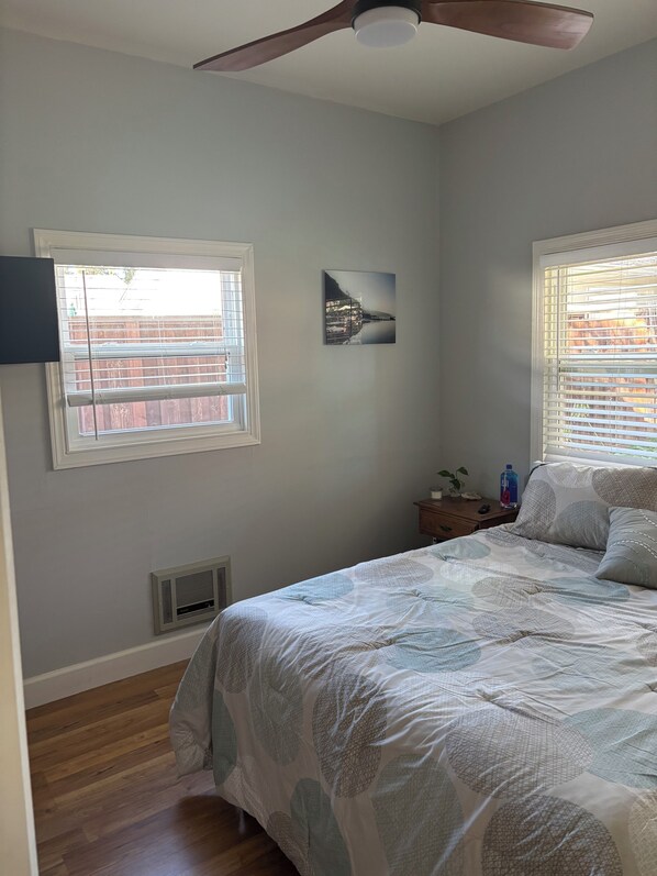 A bedroom with a bed, a ceiling fan, a window with blinds, a wall-mounted picture, and a nightstand with a lamp and a bottle.