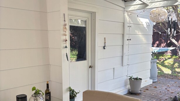 Een overdekt terras met rieten meubels, een tafel met fles en glazen, en potplanten.