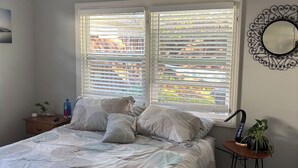 Een slaapkamer met een bed, twee ramen met zonwering, een nachtkastje met een plant, een spiegel en een plafondventilator.