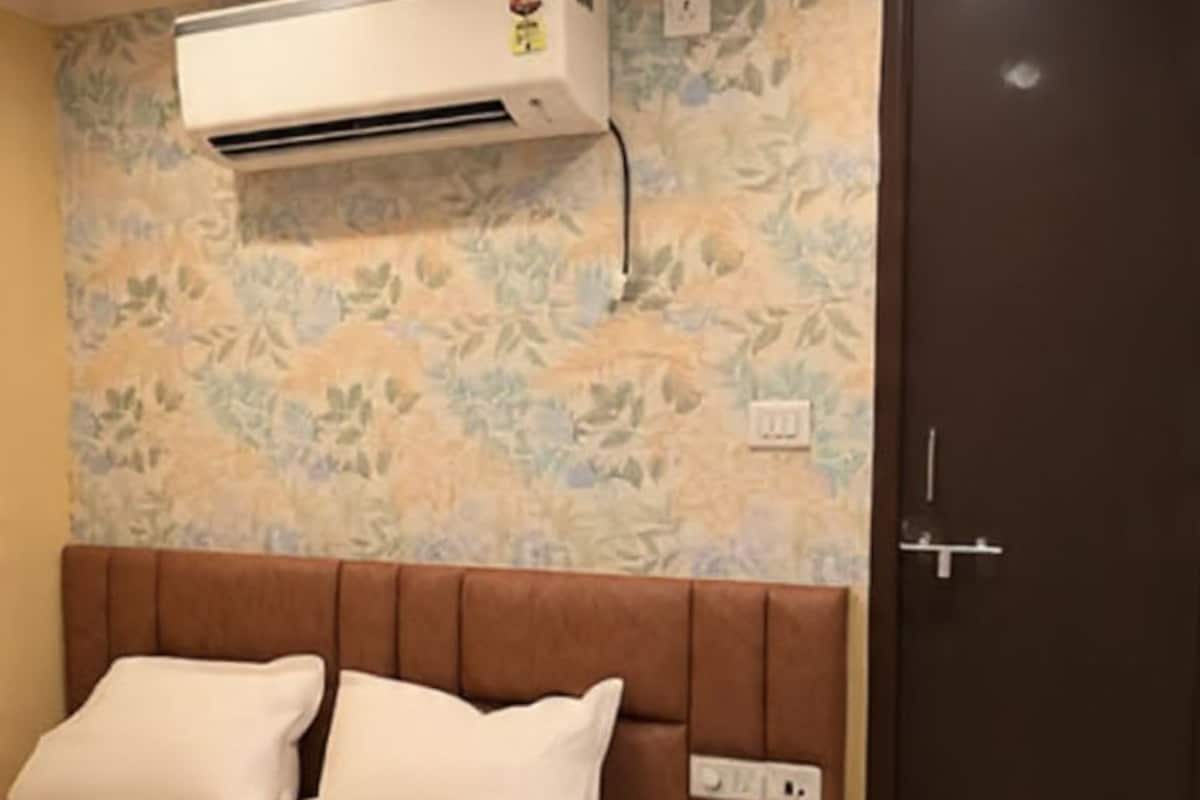 A bedroom with a bed, an air conditioning unit, a bedside table, a nightstand, and a door.