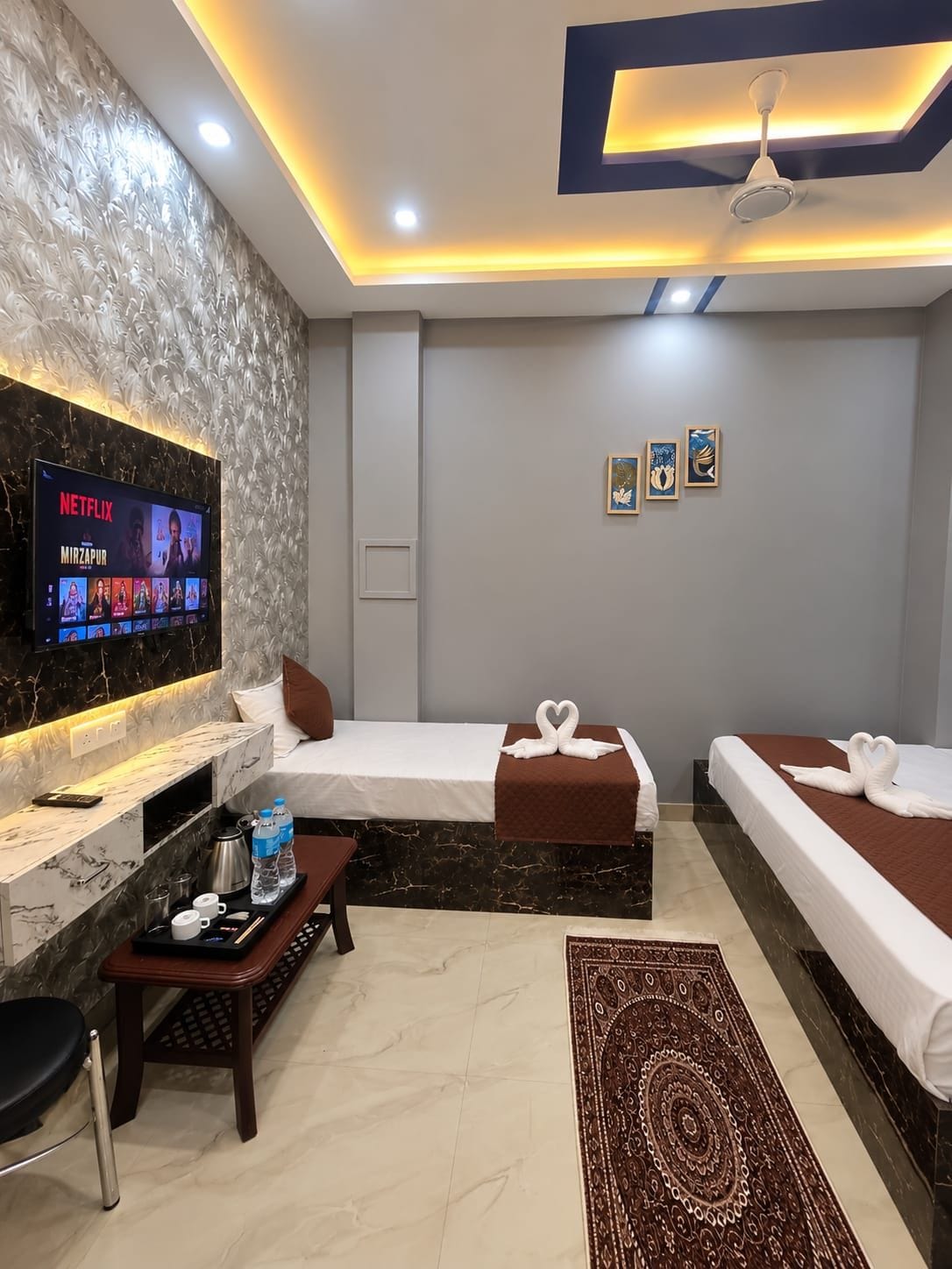 A hotel room with a flat-screen TV displaying a Netflix show, two beds with decorative pillows, a small table with a tray of amenities, and a patterned rug.