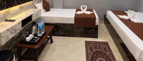 A hotel room with a flat-screen TV displaying a Netflix show, two beds with decorative pillows, a small table with a tray of amenities, and a patterned rug.