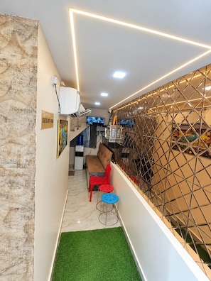 A narrow hallway with a green artificial turf floor, a red chair, a blue stool, and a white cabinet with a television. - Trinetra Homestay (Varanasi)
